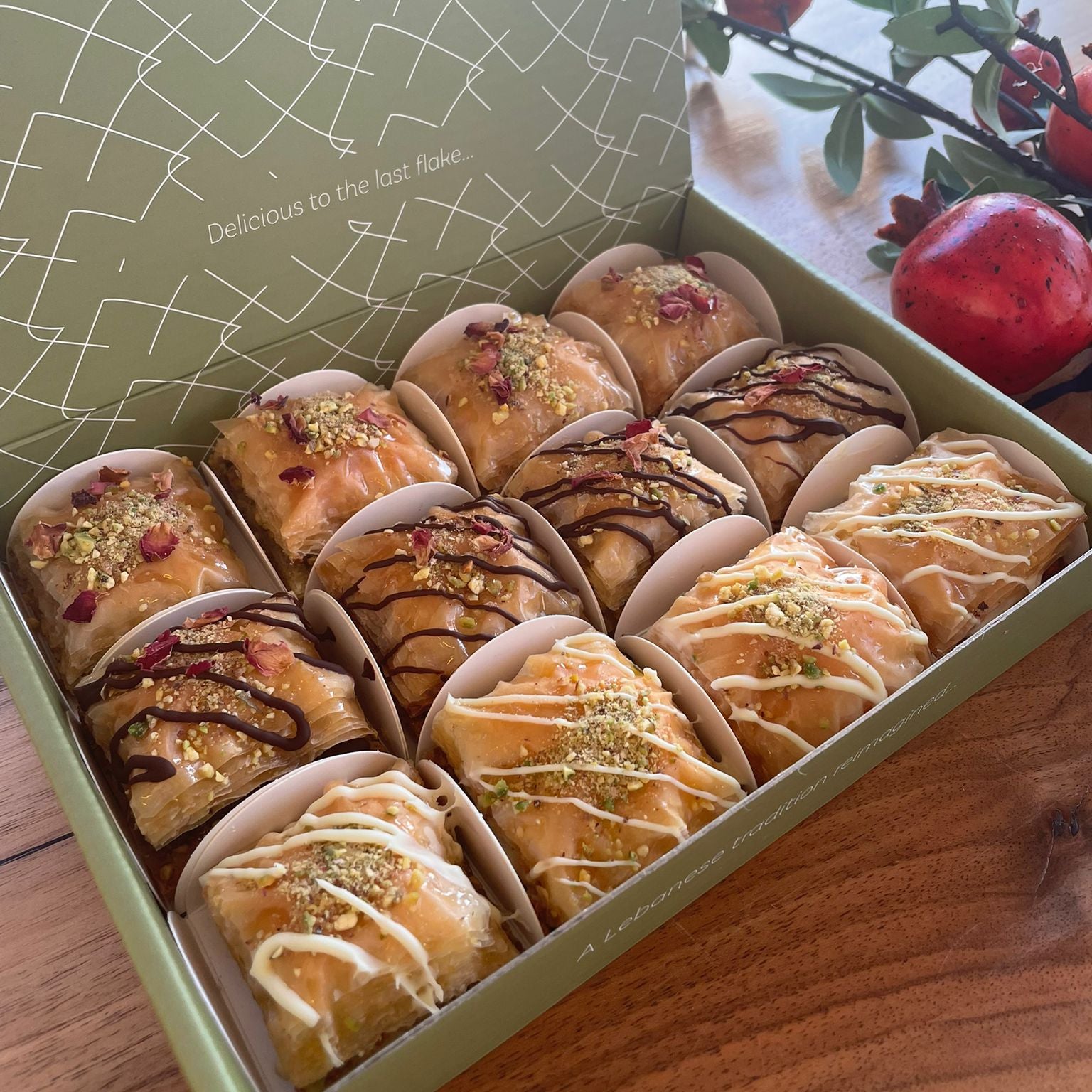 Box of assorted baklawa on a wooden surface with pomegranates in the background.